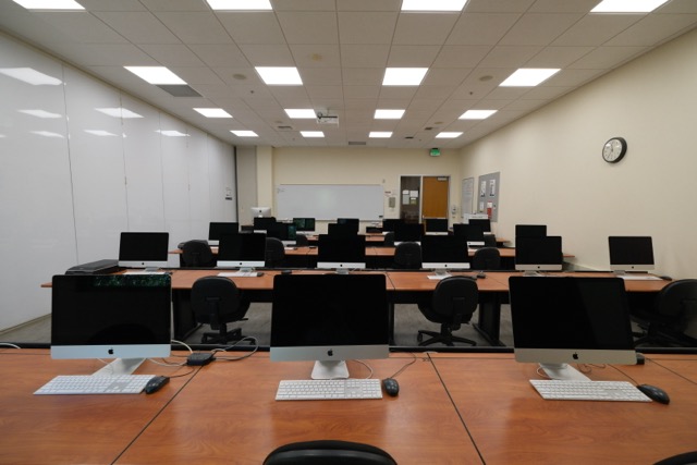 Empty computer lab with rows of desktop monitors, whiteboards, ceiling lights, clock