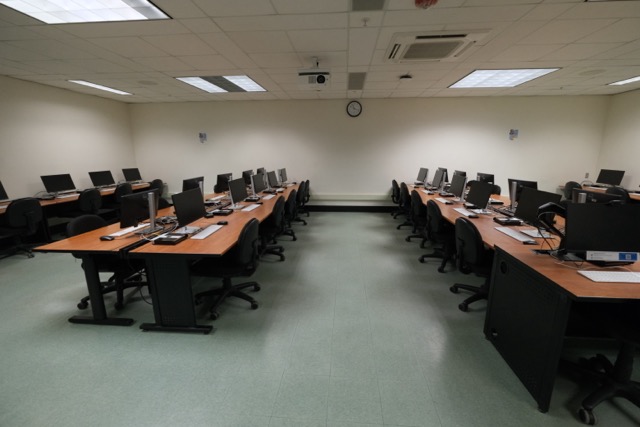 Empty computer lab with two long rows of desks, monitors and a wall clock